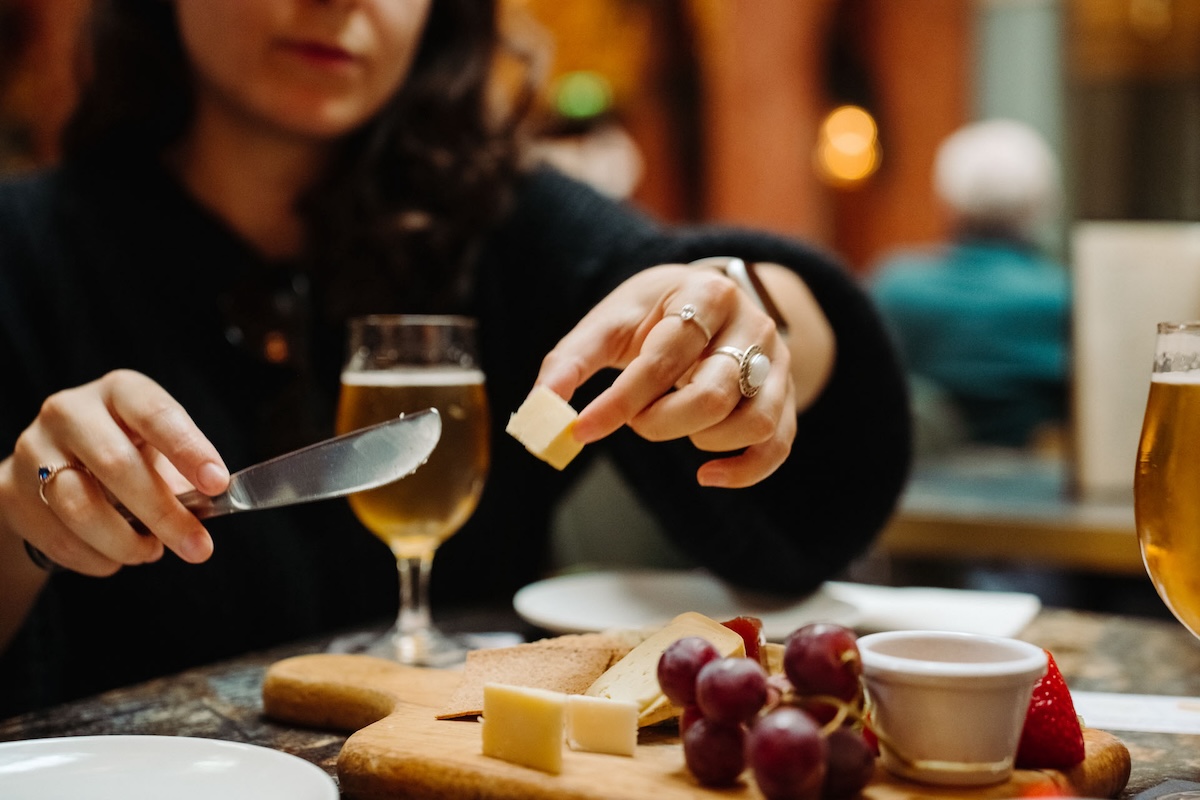 A woman slicing a piece of cheese from a charcuterie board. Along with the cheese is a variety of fruits and people drinking beer in the background.