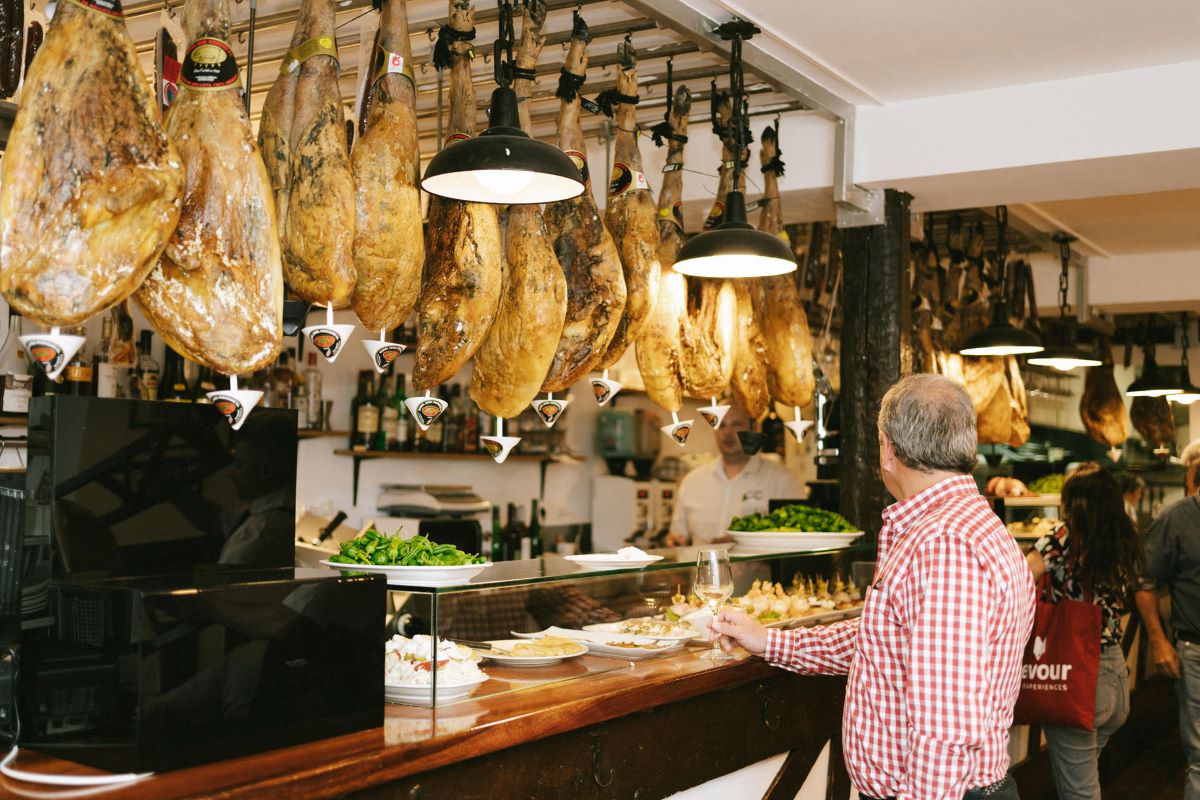 A man standing at the counter in Casa Valles. In front of him are hanging legs of jamon and pintxos in glass cases. 