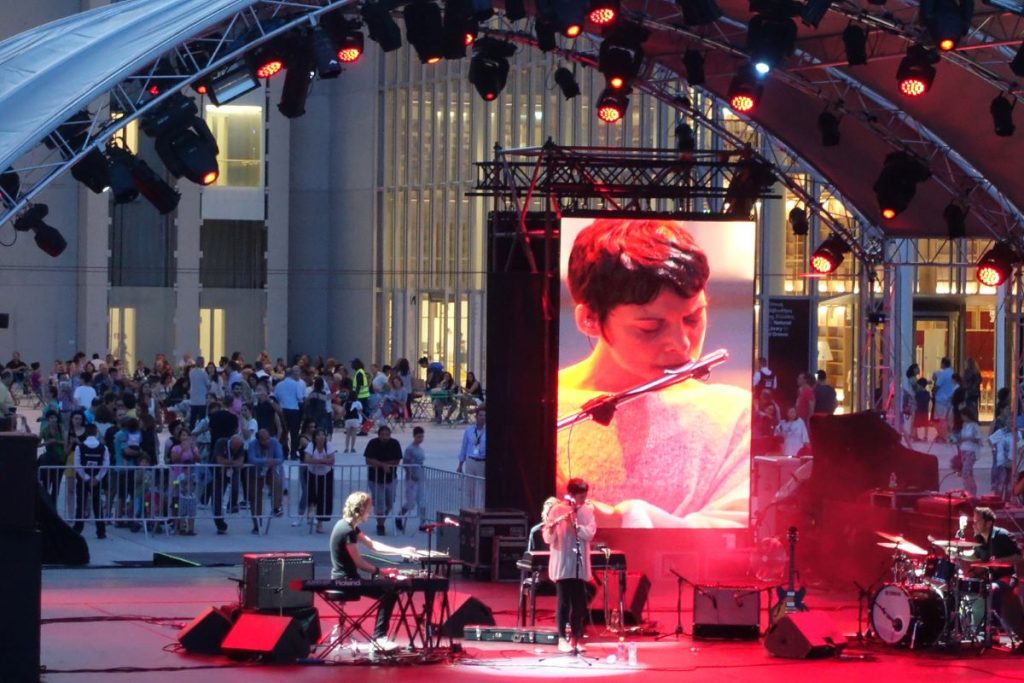 Video of a woman playing a flute on a big screen outside of Niarchos Foundation Cultural Centre.