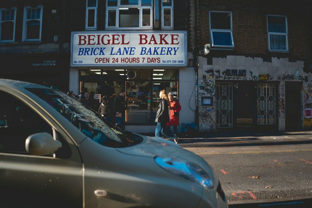 Beigel Bake storefront as a car passes in front of it and customers walk inside. 