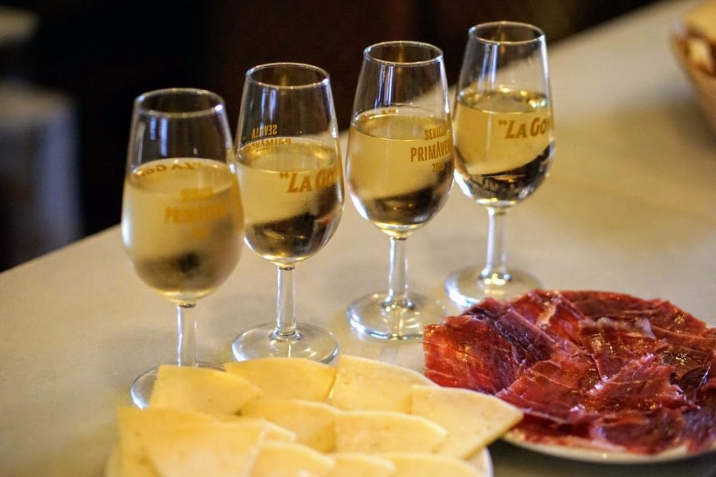 Four glasses of white sherry next to a plate of cheese and a plate of jamon during a sherry tasting in Jerez.